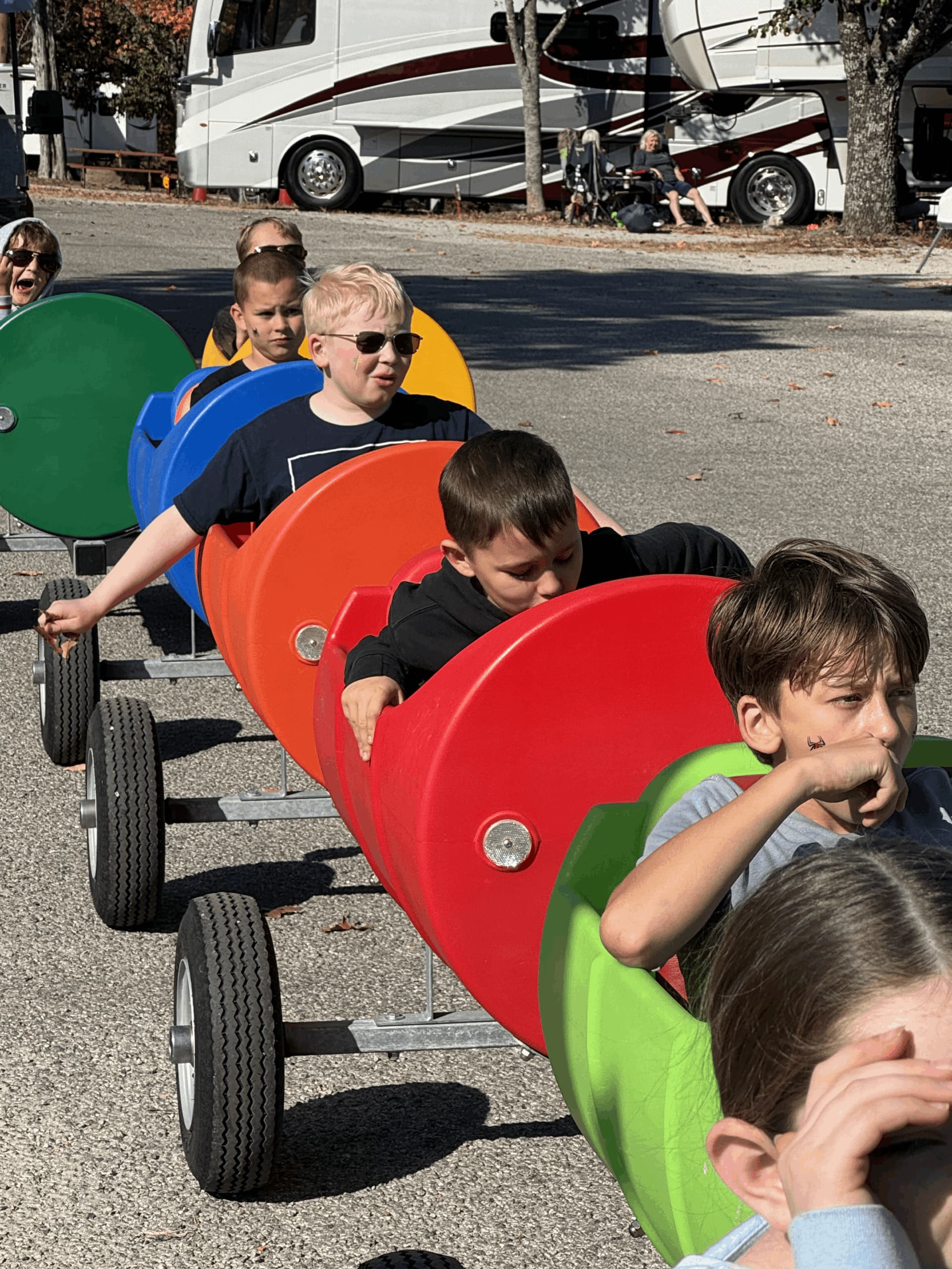 A group of children are riding on a toy train, which is red, yellow, and green